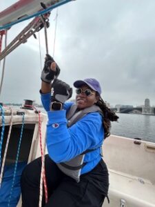 Photo of an African-American woman with coiled blocks in a turquoise hat and circular glasses. She is pulling on the van of the sales main sale. She wears a blue long sleeve T-shirt with sailing gloves marked with a brand. Black pants and a Gray life vest safely fastened. The background is an overcast with choppy water with a cityscape Of Boston behind. This appears to be a very nautical themed experience in full control of the sail.